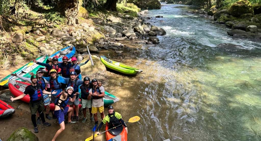 A group of people wearing safety gear stand on the shore of a river, looking up at the camera for a group photo. Their watercrafts are beached nearby.